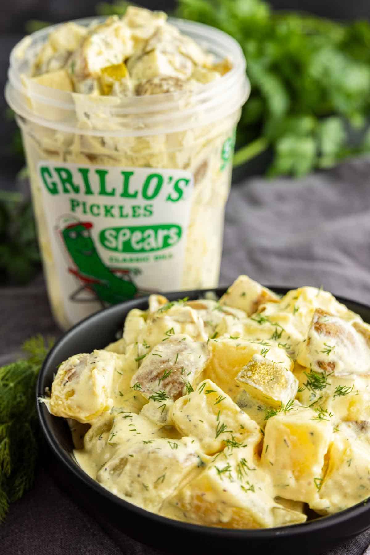A black bowl of creamy dill pickle potato salad, garnished with fresh dill, sits in front of an open container of Grillo’s Pickles Spears—an irresistible make-ahead potato salad perfect for any gathering.