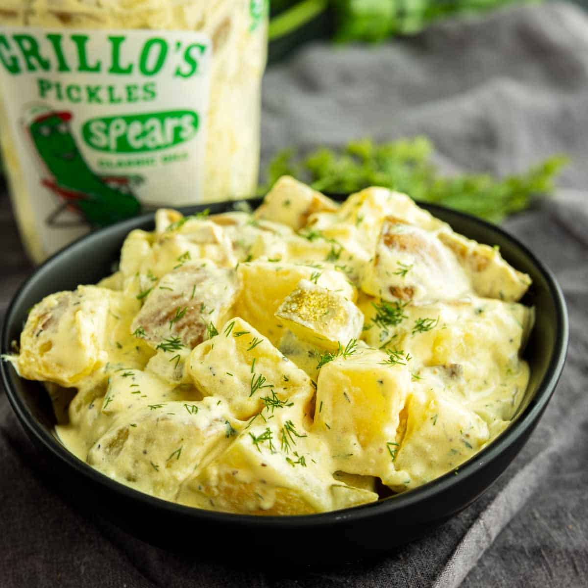 A bowl of creamy dill pickle potato salad sits in front of a container of Grillo's Pickles spears, garnished with fresh dill.