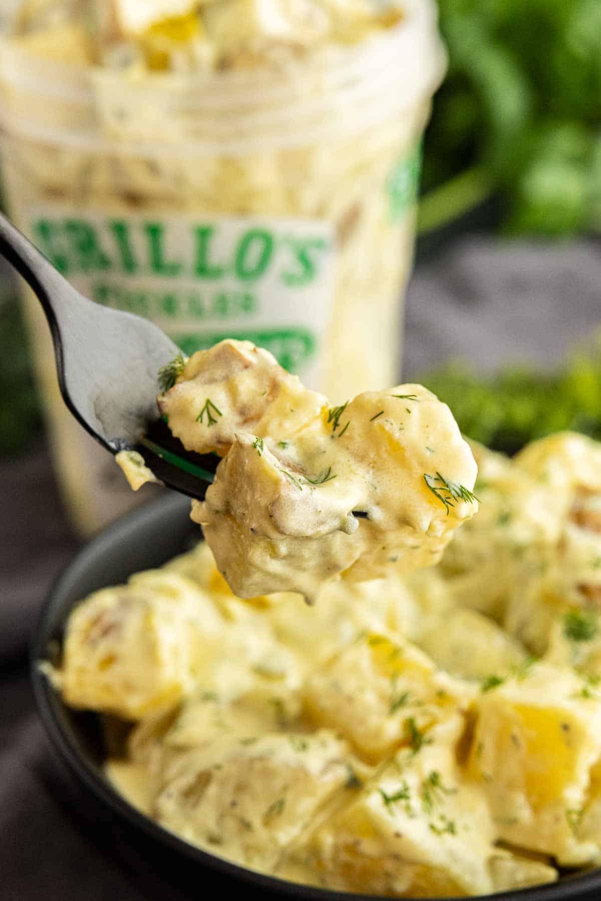 A fork holds a bite of creamy dill pickle potato salad over a bowl, with a container of Brillo’s pickles blurred in the background.