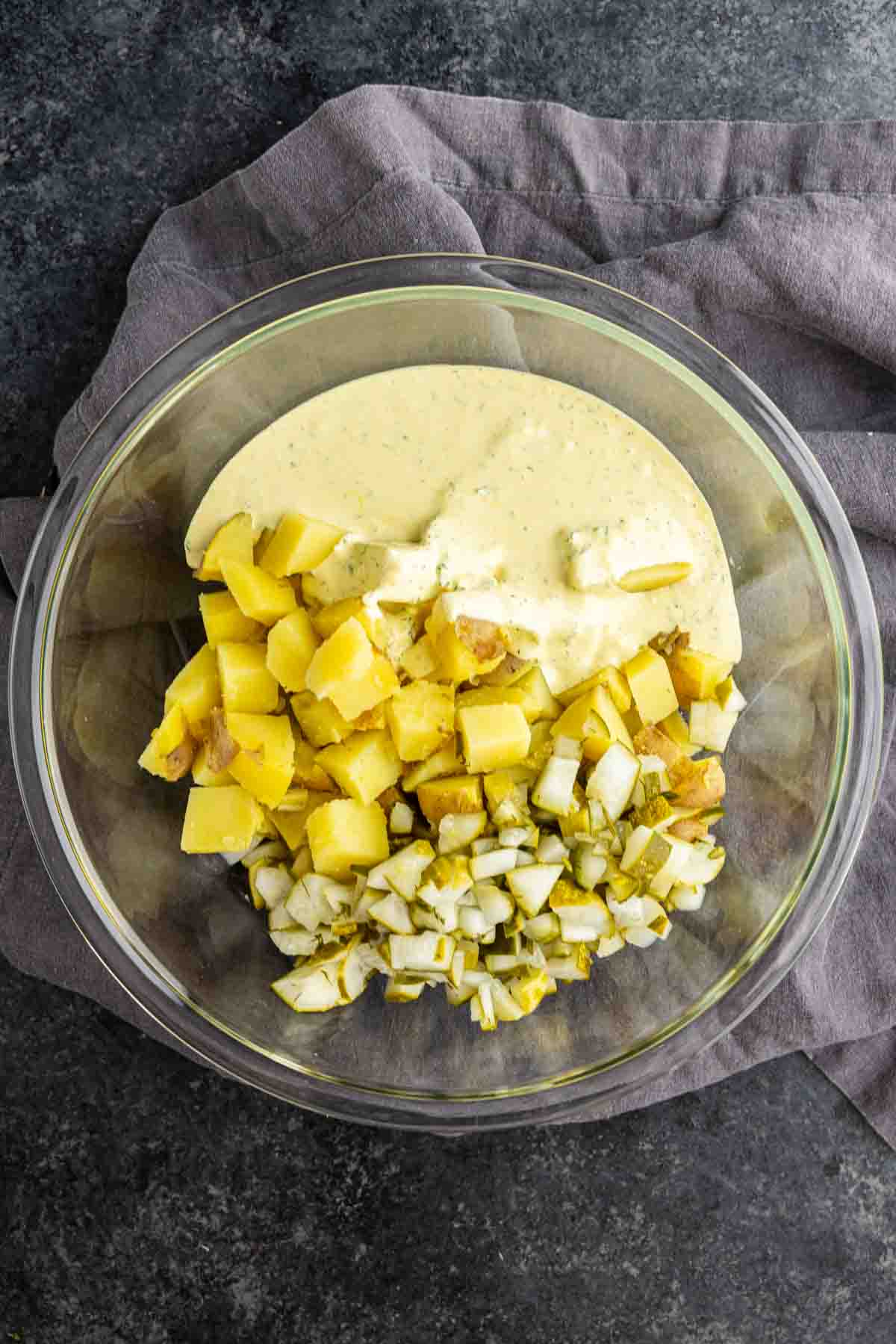 A glass bowl holds a creamy yellow pickle potato salad with chopped potatoes, diced onion, and pickles, all set atop a gray cloth.