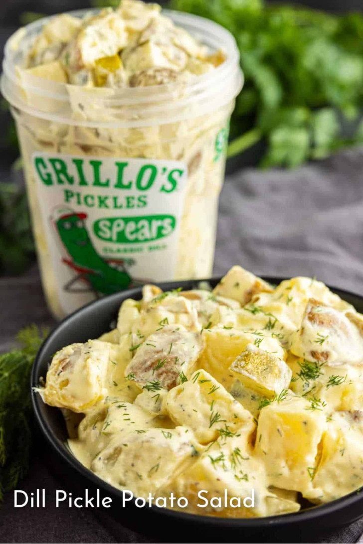 A bowl of dill pickle potato salad garnished with fresh dill sits in front of a container of Grillo’s Pickles spears.