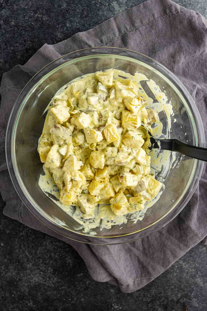 A glass bowl filled with creamy dill pickle potato salad sits on a folded gray cloth, with a black serving spoon inside the bowl.
