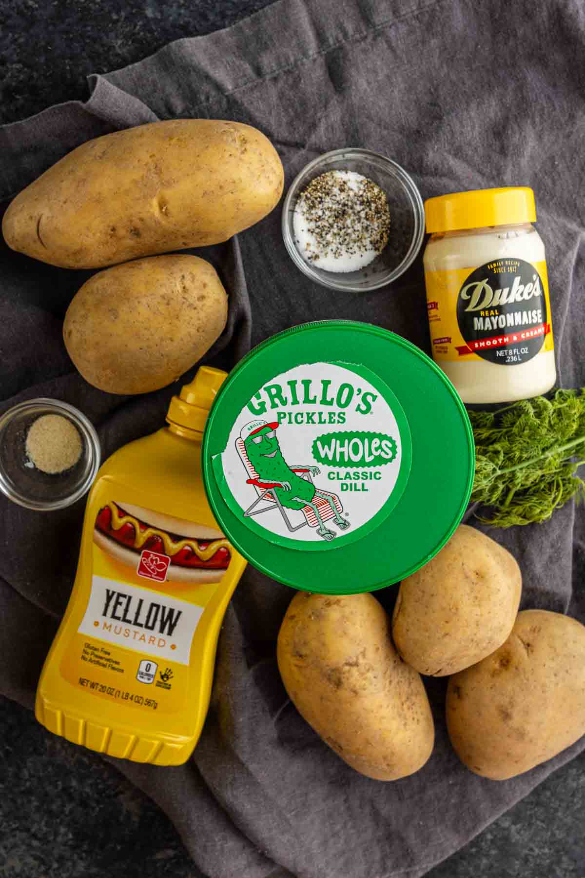 Four russet potatoes, a jar of mayonnaise, yellow mustard, dill pickles, fresh dill, ground pepper, and spices are arranged on a dark cloth—perfect for preparing a flavorful make-ahead dill pickle potato salad.