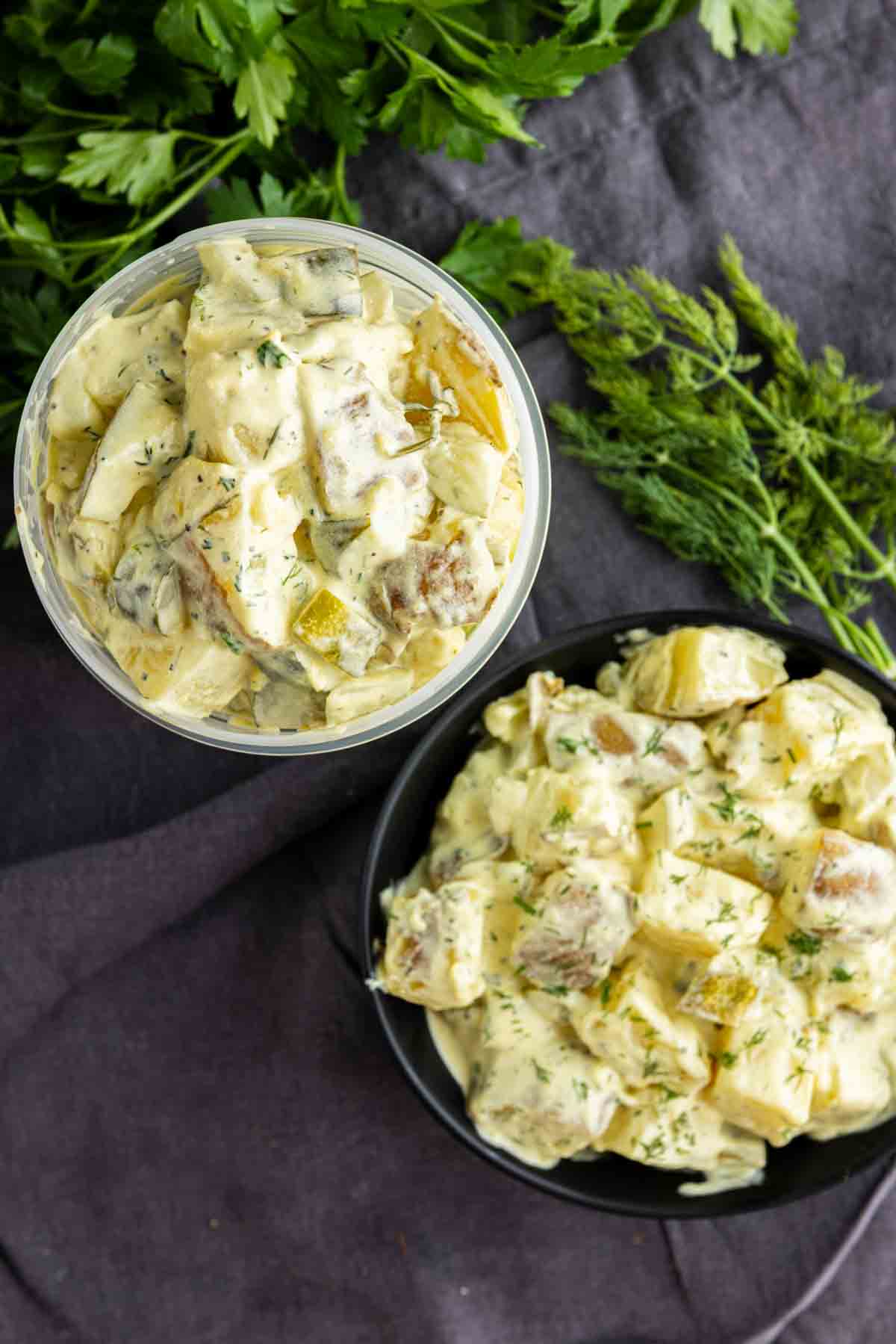 Two servings of creamy dill pickle potato salad with herbs, one in a plastic container and one in a black bowl, placed on a dark cloth next to fresh parsley and dill. This make-ahead potato salad is perfect for any occasion.