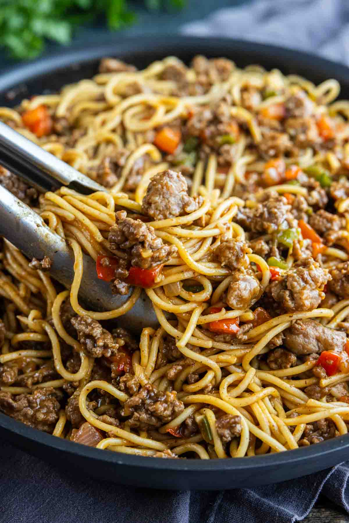 Close-up of tongs lifting stir-fried noodles with ground beef and mixed vegetables from a skillet, inspired by viral dirty spaghetti for a flavorful twist.
