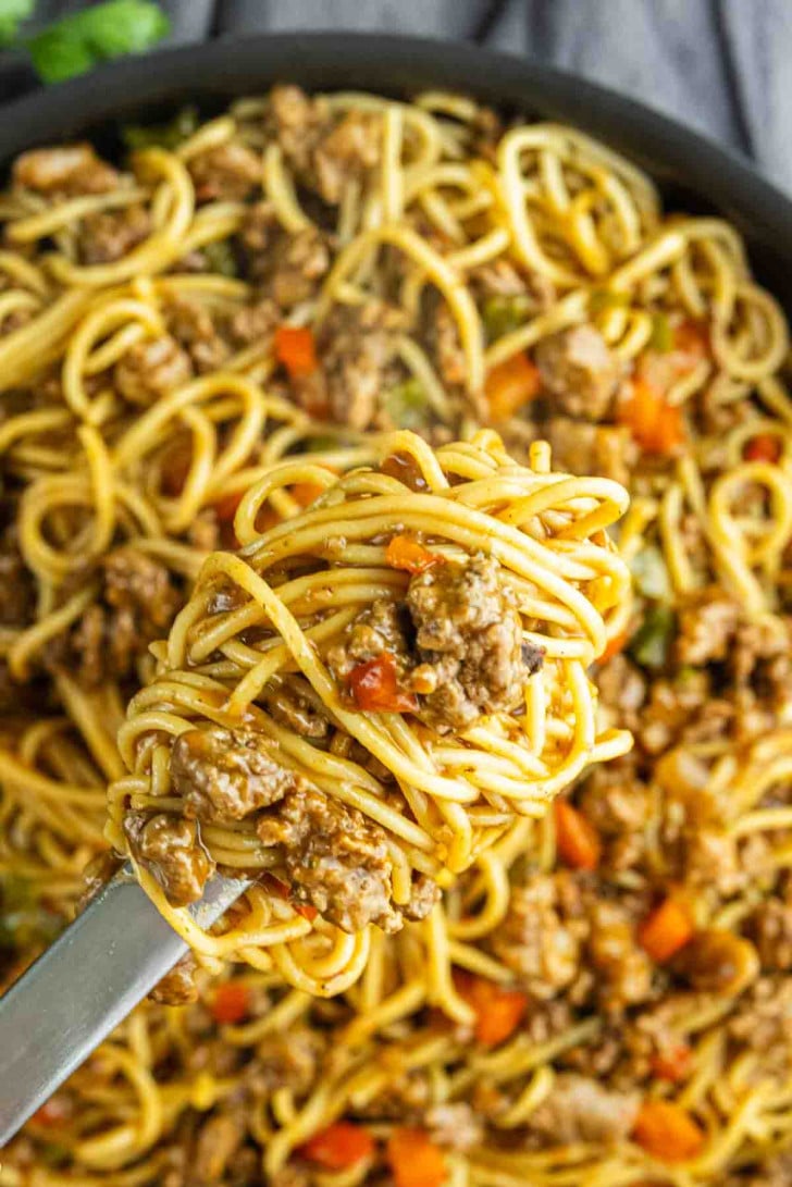 A close-up of cooked spaghetti with ground meat and diced vegetables on a fork, held above a pan filled with the same pasta dish.