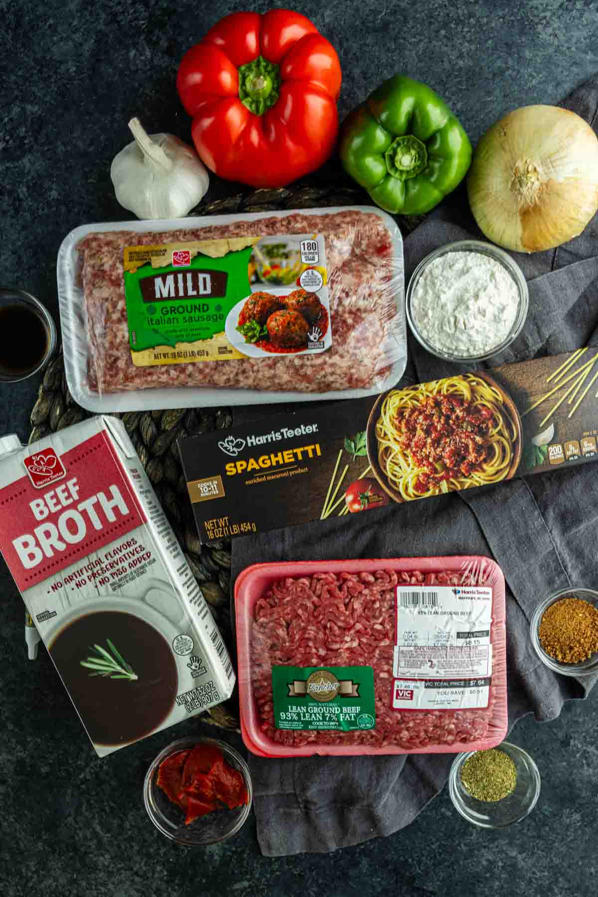 Top-down view of ingredients for Cajun spaghetti, including ground beef, Italian sausage, spaghetti pasta, bell peppers, onion, garlic, beef broth, tomato paste, and spices on a dark surface.