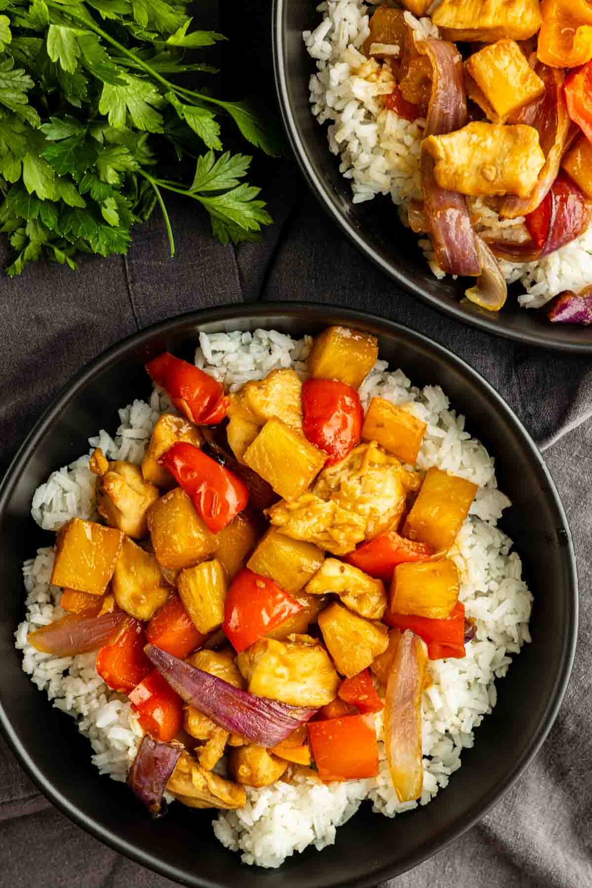 Two bowls of white rice topped with one pan Hawaiian chicken—stir-fried with red bell peppers, pineapple chunks, and red onions—sit on a dark tablecloth beside fresh parsley. An easy weeknight dinner that's bursting with tropical flavor.