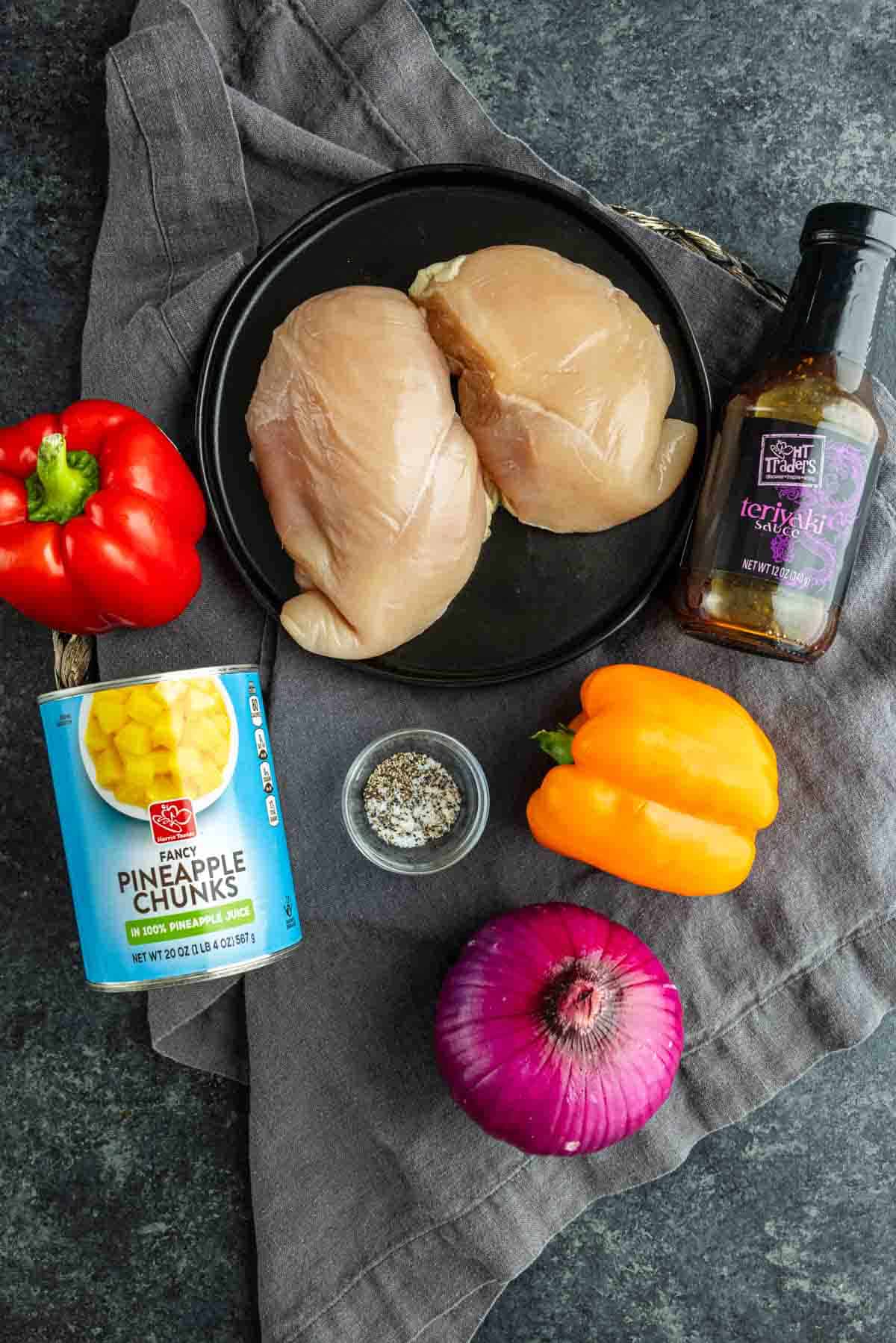 Raw chicken breasts on a black plate, surrounded by red and yellow bell peppers, red onion, pineapple chunks, seasoning blend, and teriyaki sauce—perfect for an easy weeknight dinner like sheet pan Hawaiian chicken.