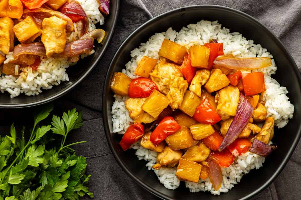 A bowl of white rice topped with sweet and sour chicken, bell peppers, pineapple chunks, and red onion—this one pan Hawaiian chicken makes for an easy meal. Another partial bowl and fresh parsley are next to it.