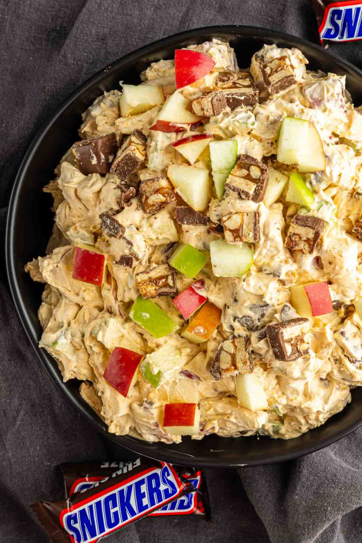 A bowl filled with chopped Snickers bars, red and green apple pieces, and creamy dessert mixture, with Snickers wrappers beside the bowl.