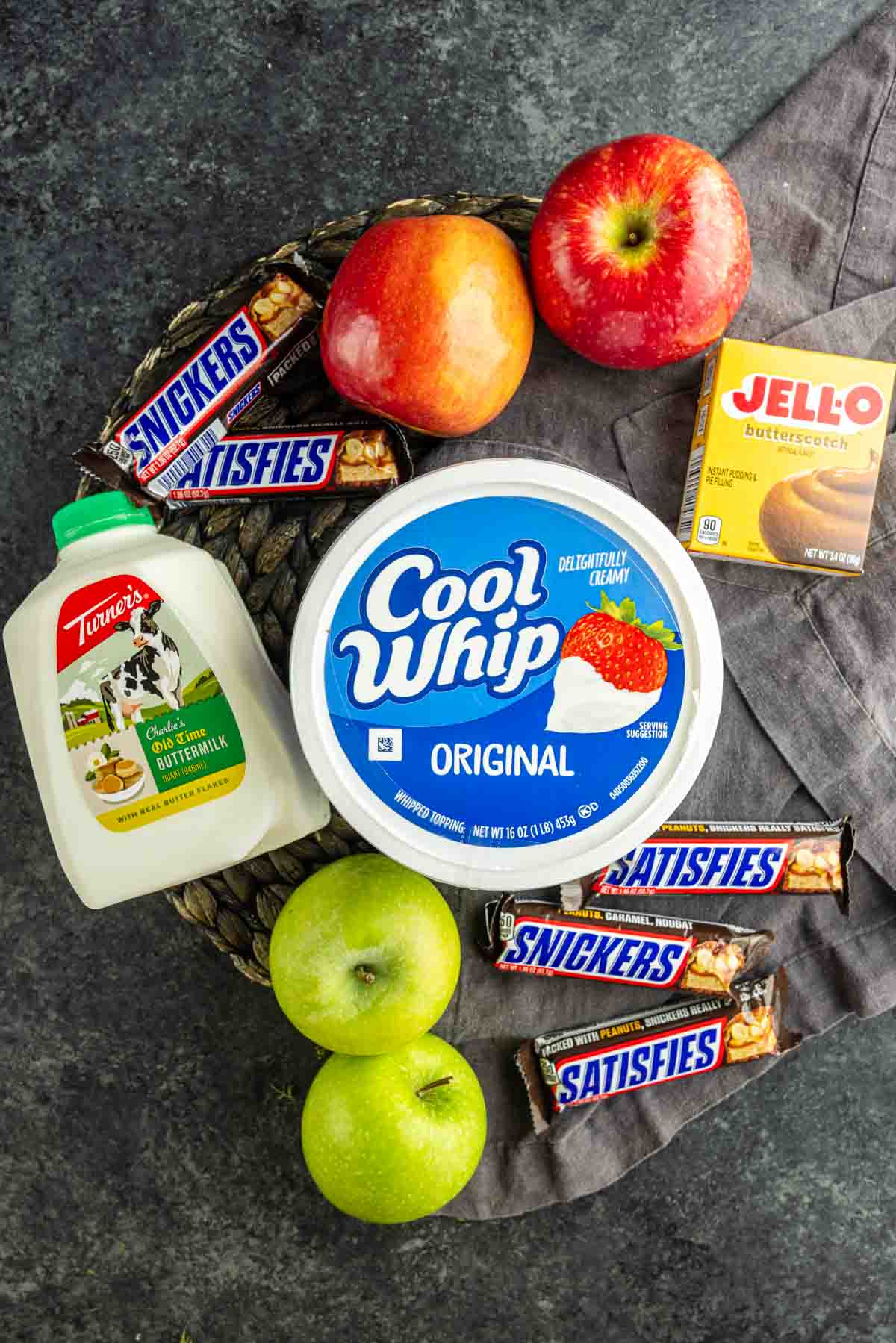 A classic Snickers Salad recipe: four Snickers bars, two red apples, two green apples, a carton of buttermilk, Cool Whip, and a box of Jello butterscotch pudding mix on a dark surface—everything you need for an easy Snickers Salad.
