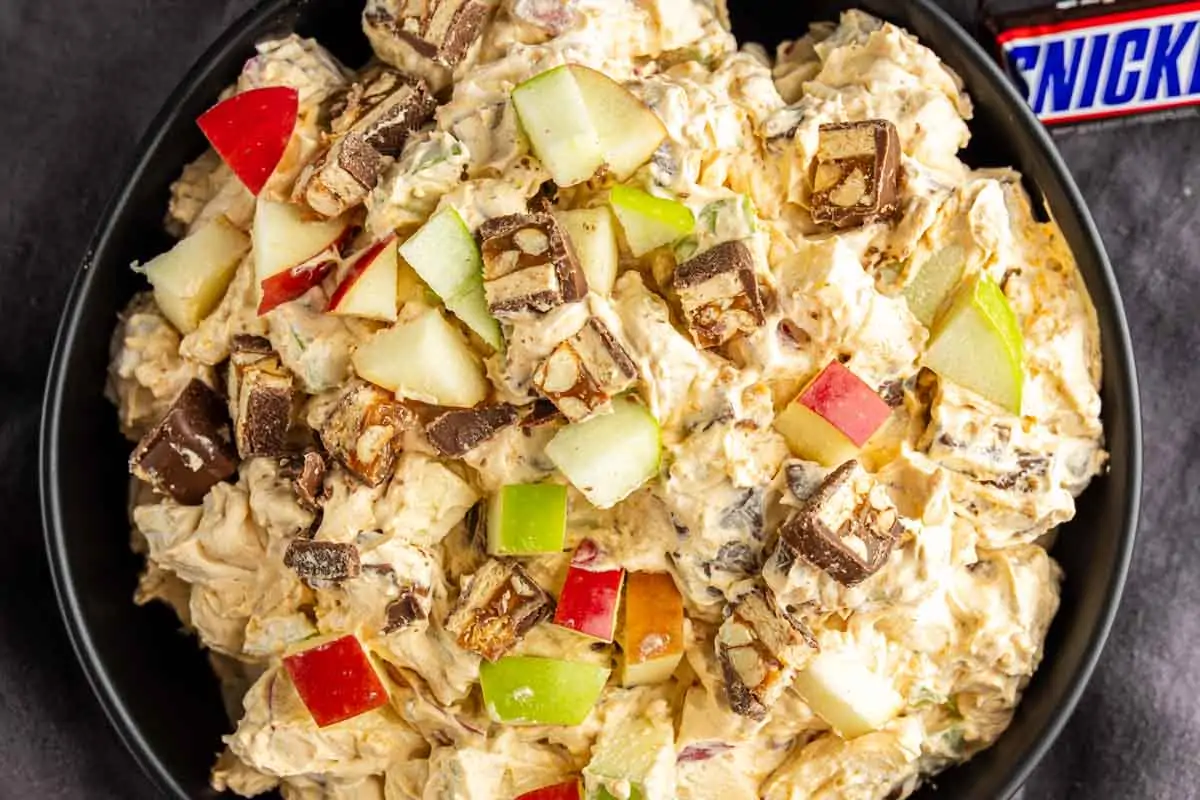 A bowl of creamy apple Snickers salad topped with chopped Snickers bars and red and green apple pieces.