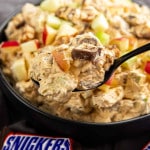 A spoonful of creamy Snicker Apple Salad with chopped apples and pieces of Snickers bar held above a bowl of the same salad; a Snickers wrapper is visible in the foreground.