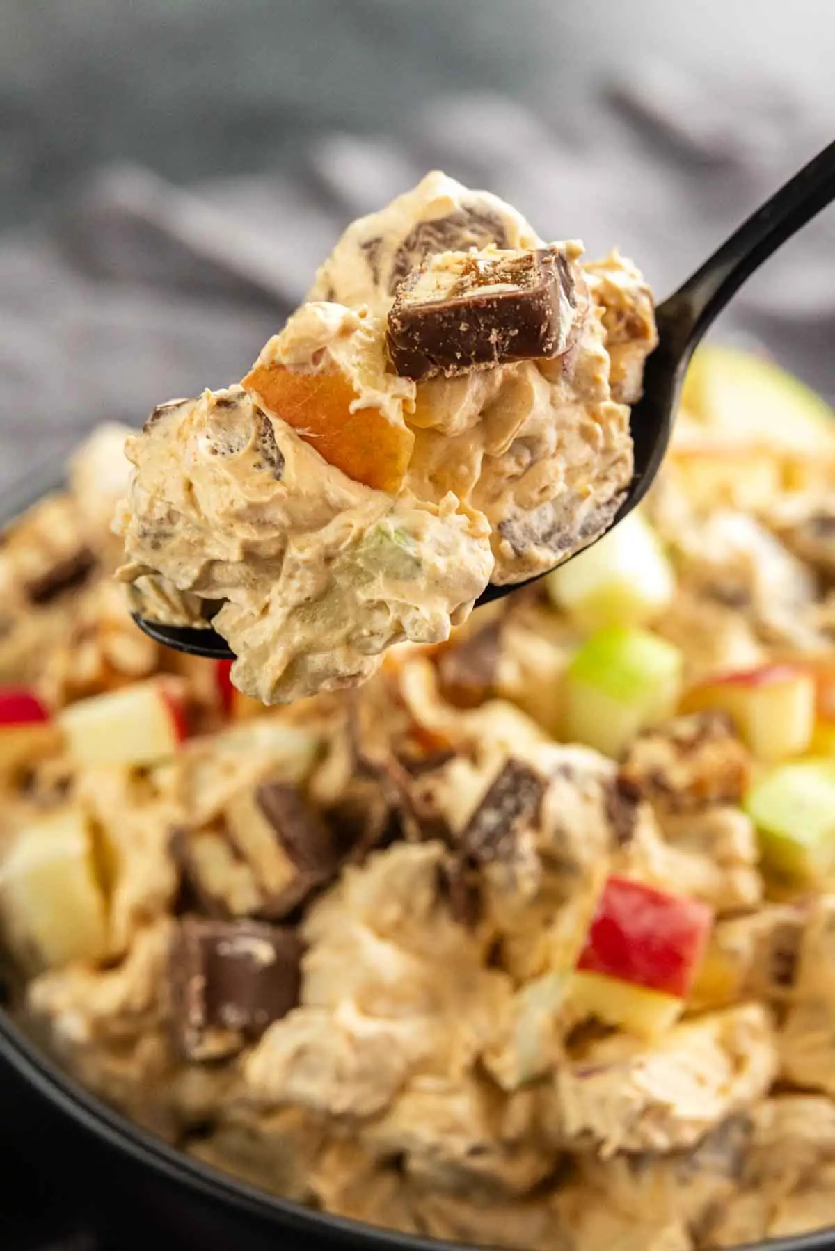 A close-up of a spoonful of creamy Snickers Apple Salad with chopped apples and chocolate pieces, held above a bowl filled with the same mixture.