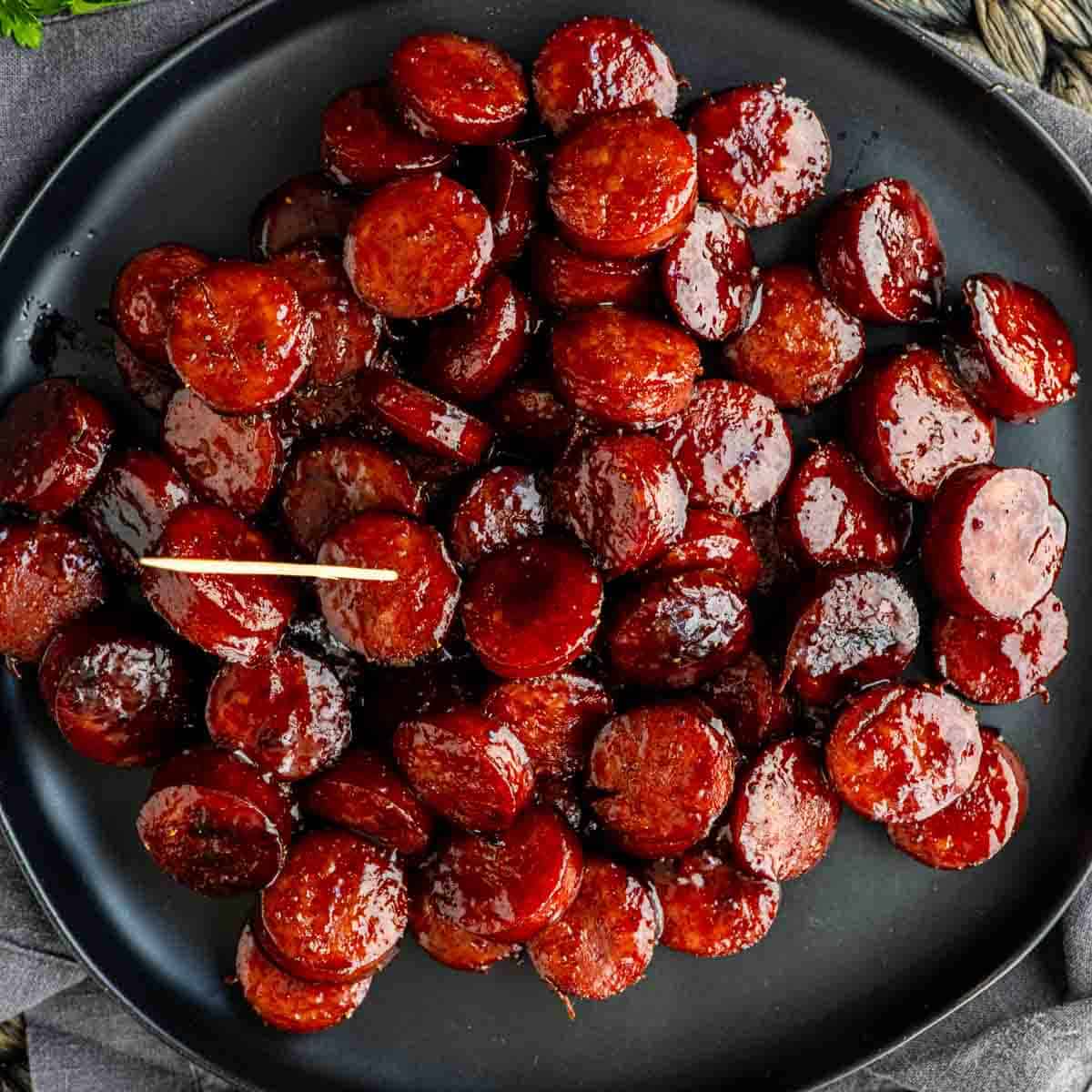 A black plate filled with glazed BBQ kielbasa bites is shown, each sliced sausage round glistening and one piece topped with a toothpick.