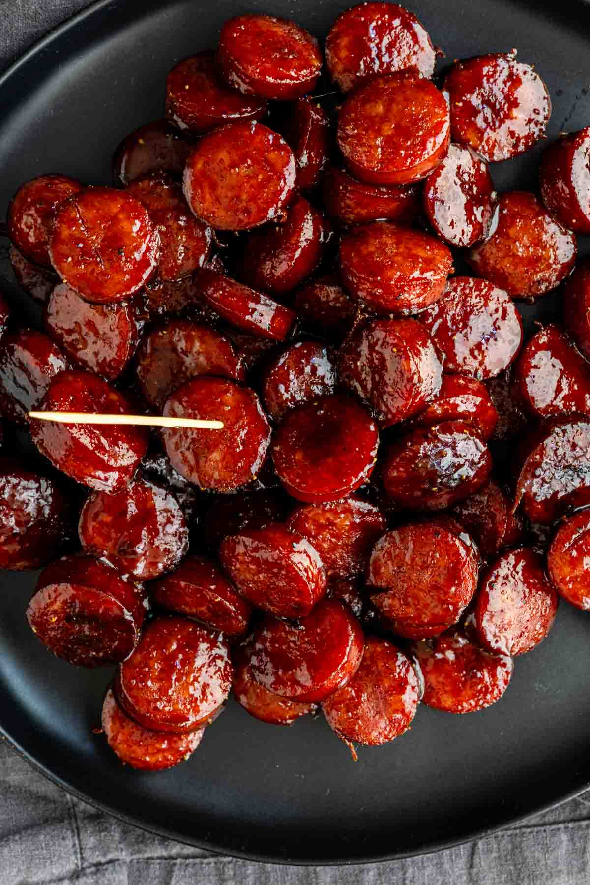 Sliced sausage pieces coated in a glossy Barbecue Brown Sugar Kielbasa sauce are arranged on a black plate, with a toothpick placed among them.