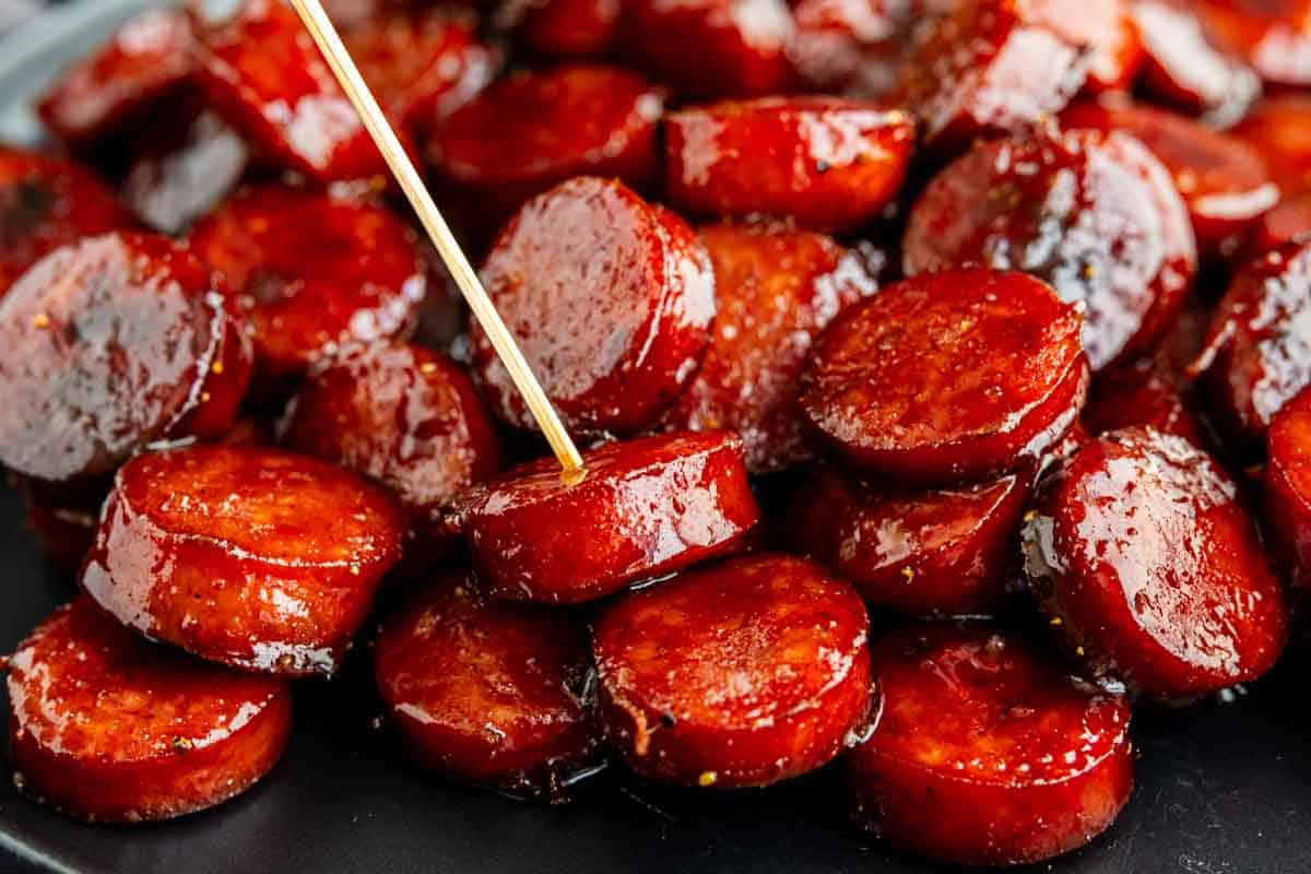 A close-up of BBQ kielbasa bites stacked on a plate, glazed to perfection, with a toothpick inserted into one of the slices.