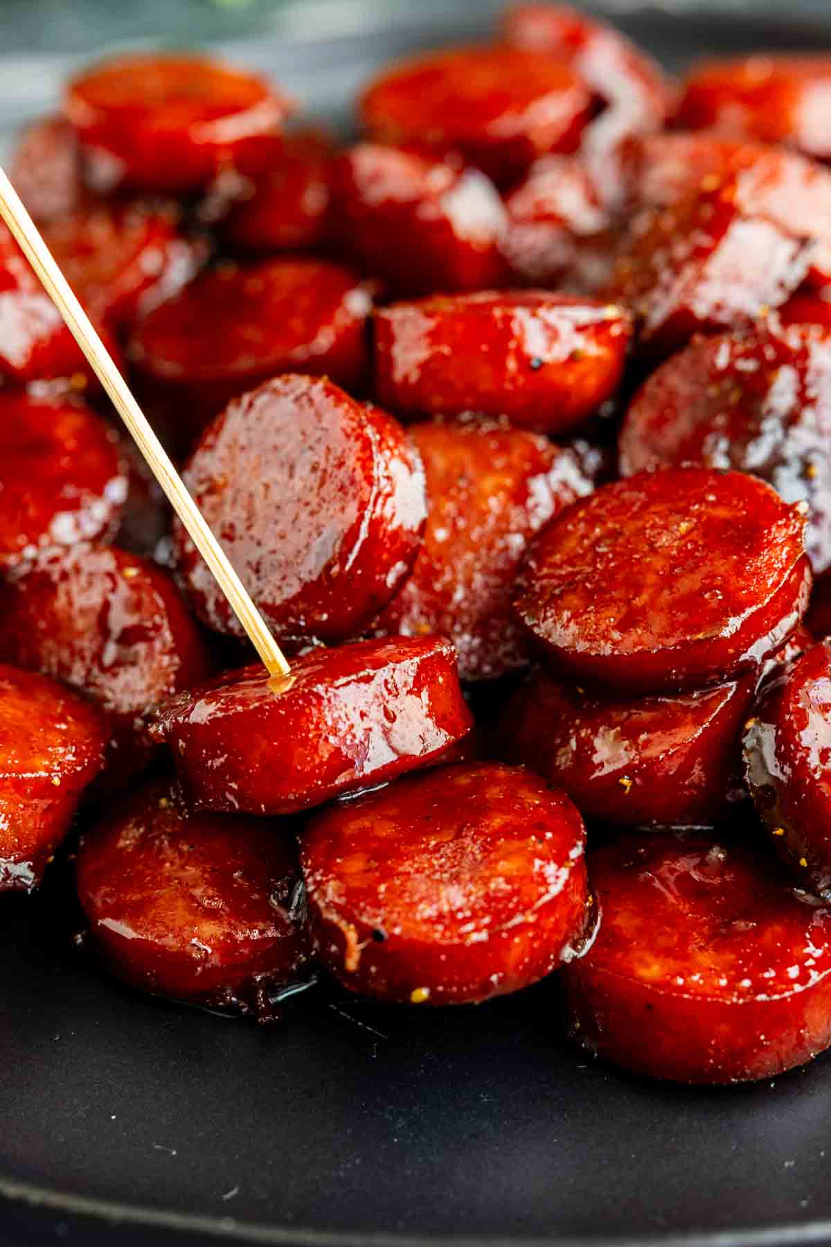 Close-up of Barbecue Brown Sugar Kielbasa bites on a black plate, with a toothpick inserted into one of the glazed sausage slices—an irresistible kielbasa appetizer recipe.