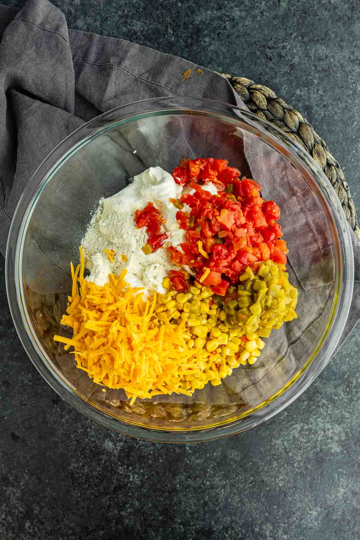 A glass bowl filled with shredded cheddar cheese, chopped green chilies, diced tomatoes, sour cream, and a dry seasoning mix sits on a dark surface with a gray cloth—an irresistible viral boat dip ready to be served.