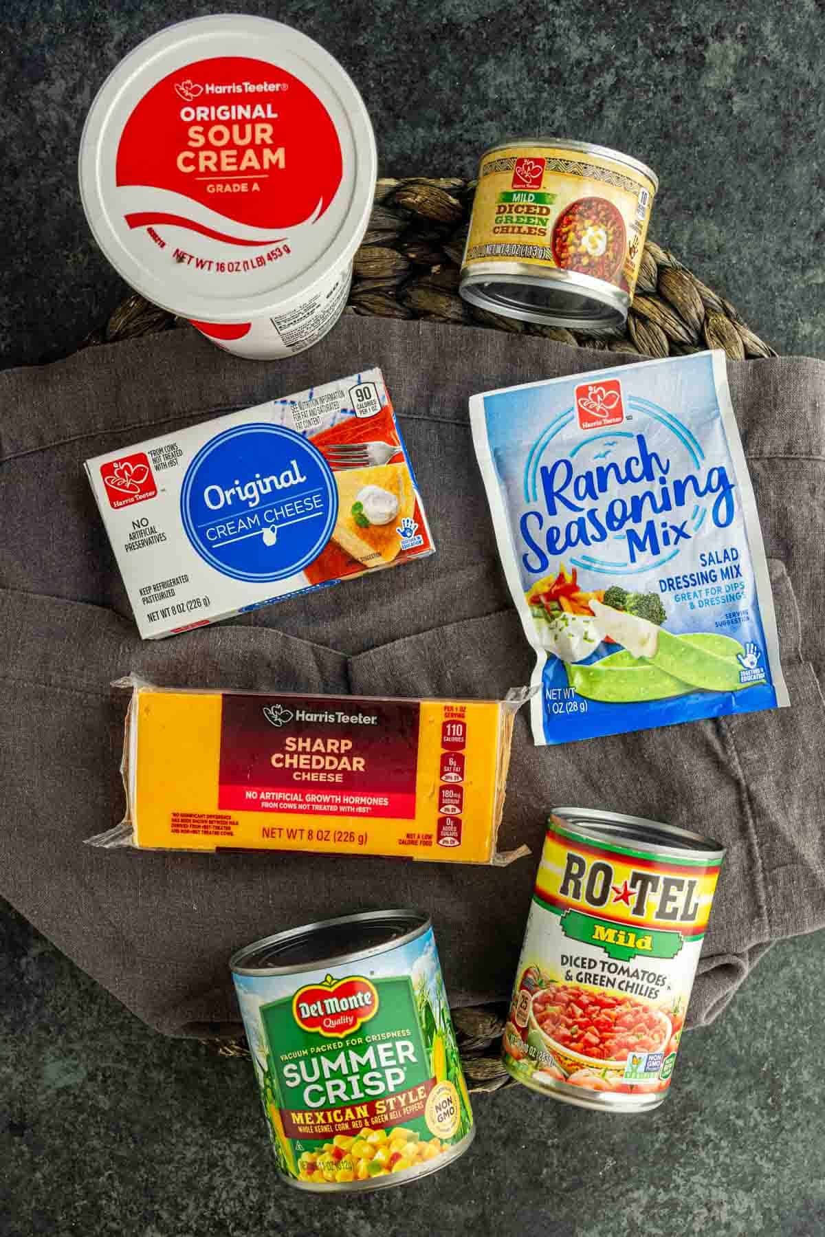 A variety of packaged food items, perfect for making cold corn dip or the viral boat dip, including sour cream, cream cheese, sharp cheddar cheese, ranch seasoning mix, summer crisp corn, and diced tomatoes with green chilies on a gray surface.