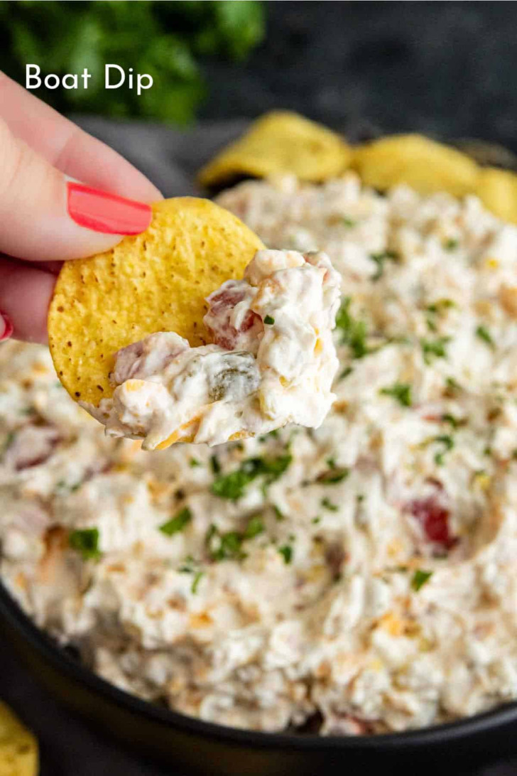 A hand holding a tortilla chip with creamy dip above a bowl filled with the same dip, garnished with chopped herbs. The word "Boat Dip" is written in the image.