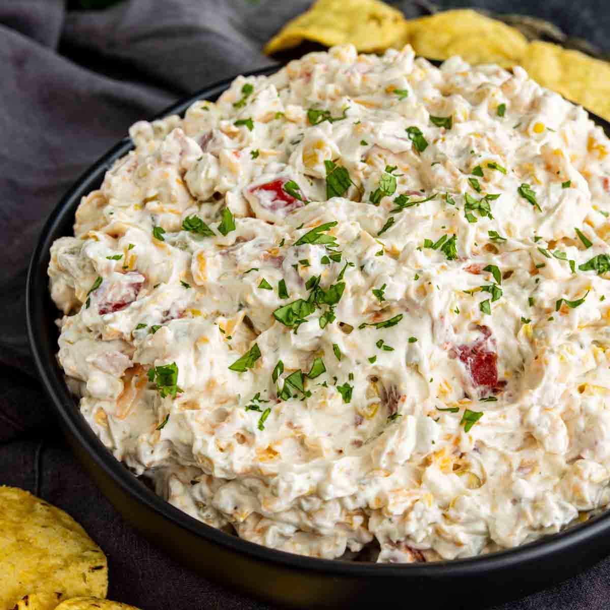 A black bowl filled with creamy cold corn dip containing chopped tomatoes and herbs, garnished with parsley, surrounded by tortilla chips.