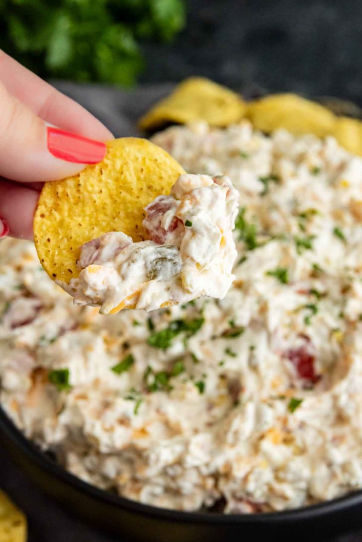 A close-up of a hand holding a tortilla chip with creamy Rotel ranch dip, in front of a bowl filled with the same dip topped with chopped herbs.