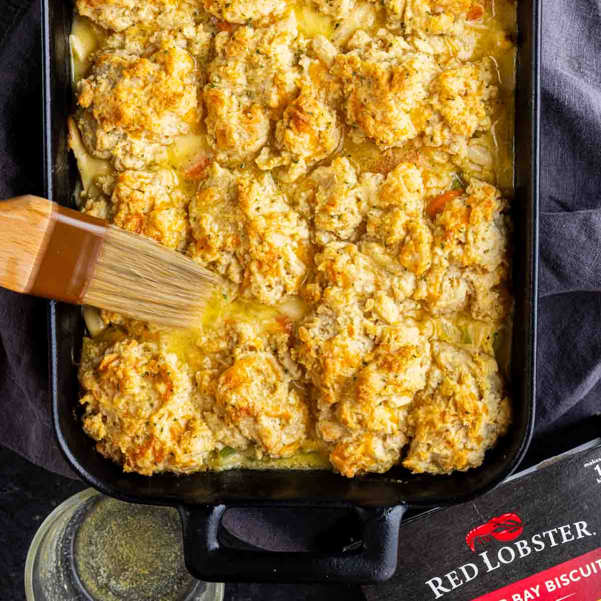 A baking dish filled with golden-brown Cheddar Bay Biscuit Chicken Casserole is being brushed with melted butter; a Red Lobster biscuit mix box is partially visible.