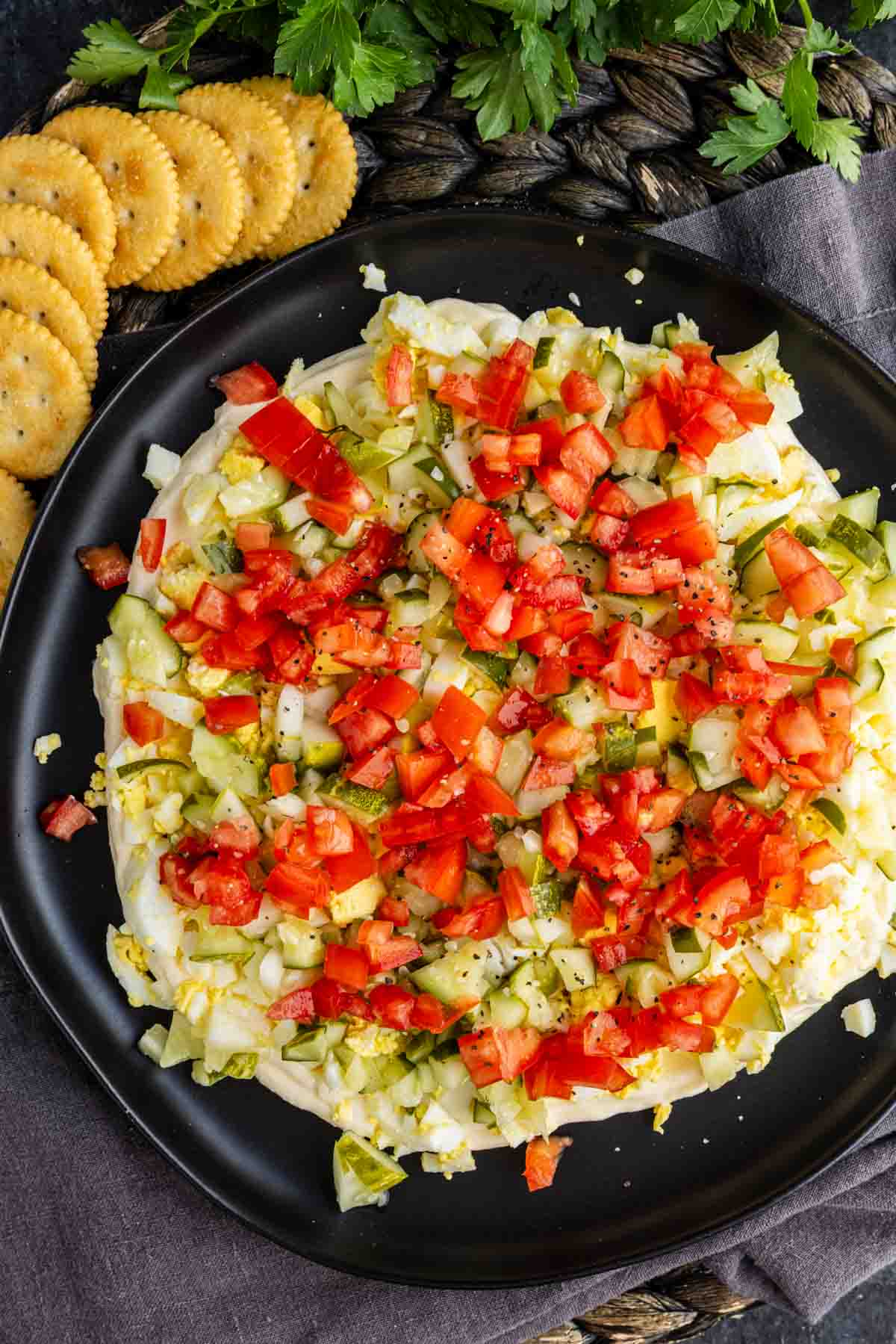 A black plate holds layered egg salad dip covered with chopped tomatoes, pickles, and onions, surrounded by round crackers. Parsley is partially visible at the top of the image.