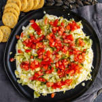 A round plate topped with a layered egg salad dip featuring diced pickles, onions, and tomatoes on a creamy base, served with crackers on the side.