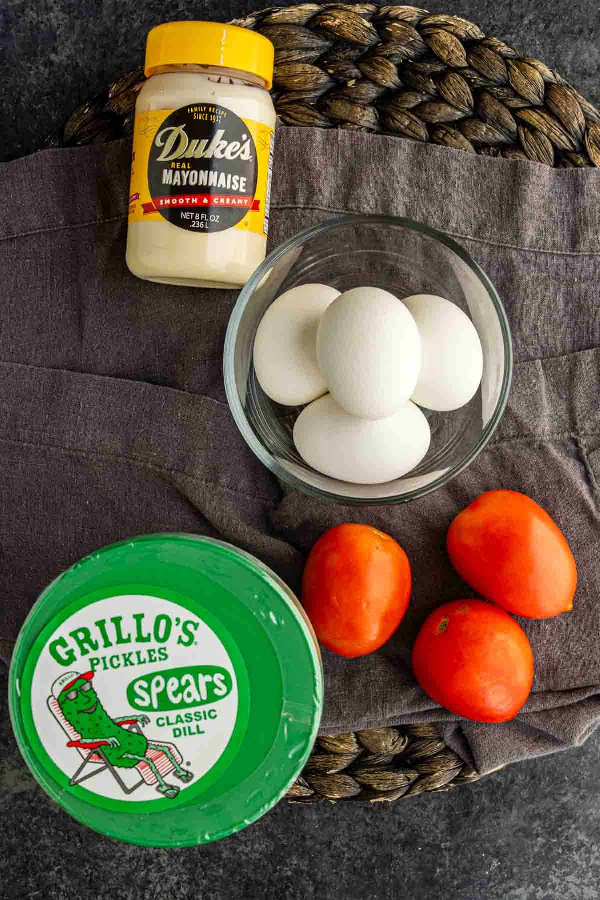 A jar of mayonnaise, a bowl of four eggs, three Roma tomatoes, and a container of dill pickles arranged on a dark cloth and woven mat—perfect ingredients for a delicious layered egg salad dip.