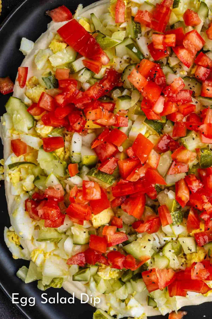 Close-up of an egg salad dip topped with chopped tomatoes, pickles, and onions on a black plate.