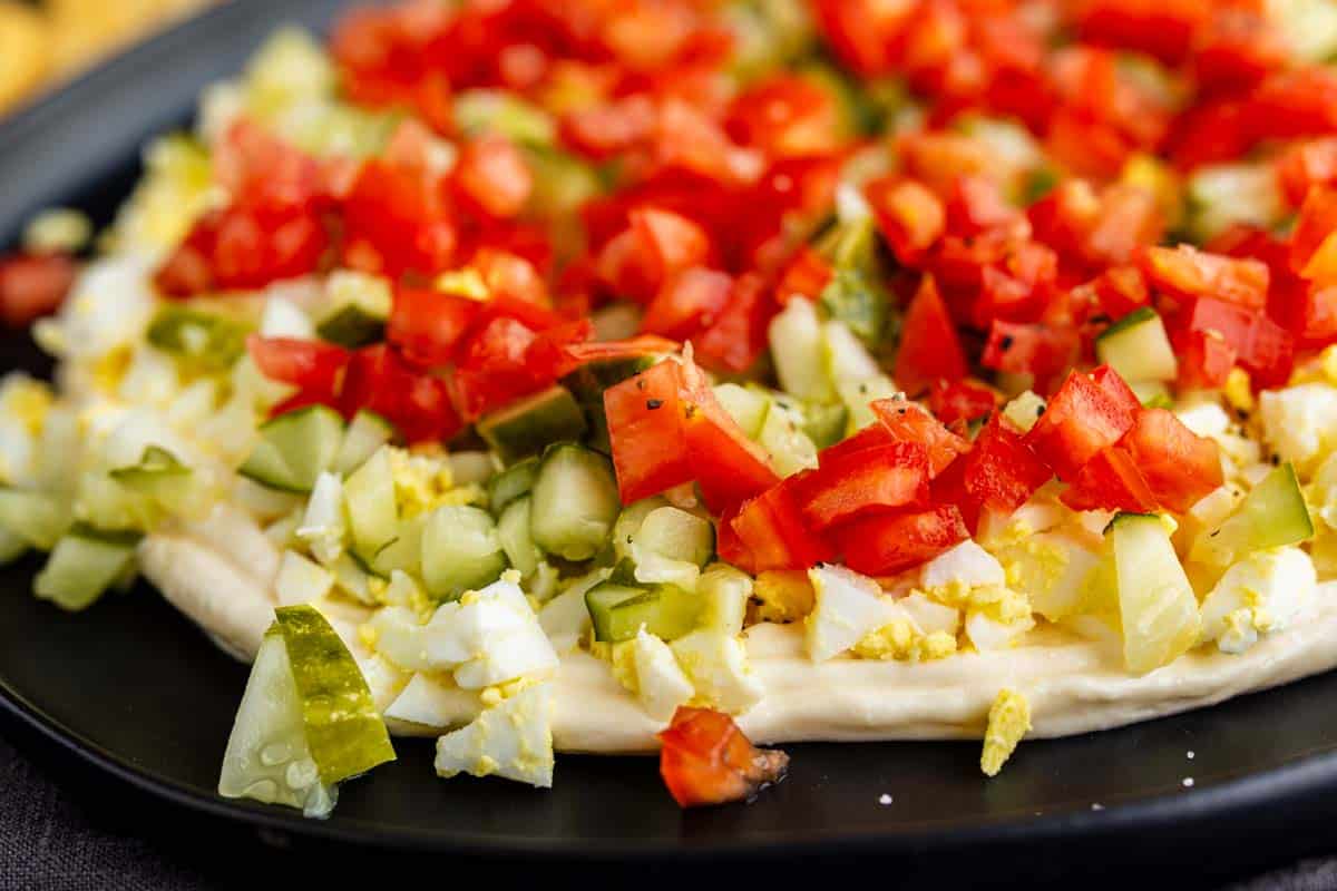 Close-up of a flatbread topped with chopped hard-boiled eggs, diced pickles, and diced tomatoes on a black plate—an easy twist on the popular TikTok egg salad dip.