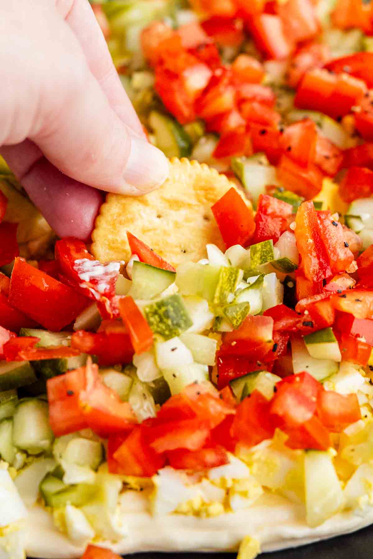 A hand dips a round cracker into a layered egg salad dip, topped with diced tomatoes, pickles, onions, and peppers on a creamy base.