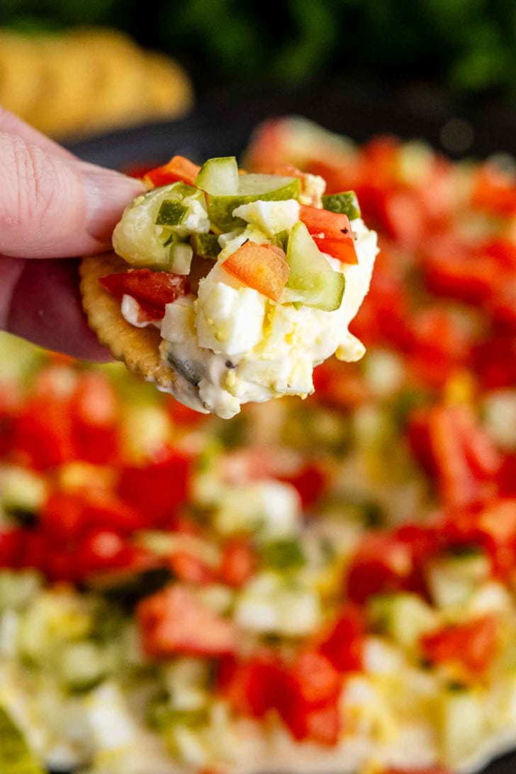 A hand holding a cracker topped with a creamy spread, diced cucumber, tomato, and green onions, with more of the dip visible in the background.