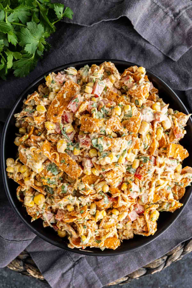A bowl of salad made with corn, diced vegetables, shredded cheese, and crushed corn chips on a dark cloth background.