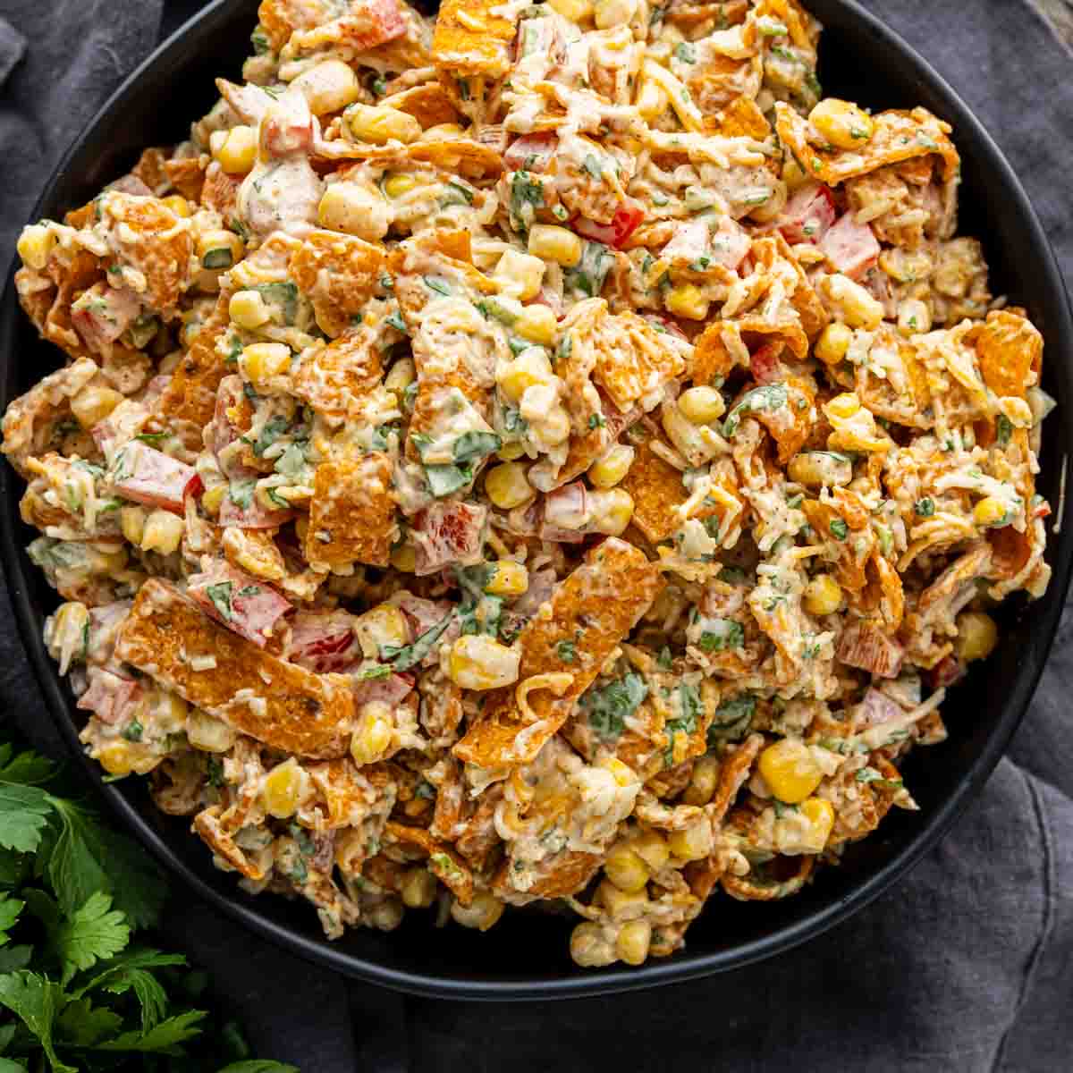 A bowl filled with a creamy corn chip salad made of corn, chopped vegetables, herbs, and crushed tortilla chips mixed together.