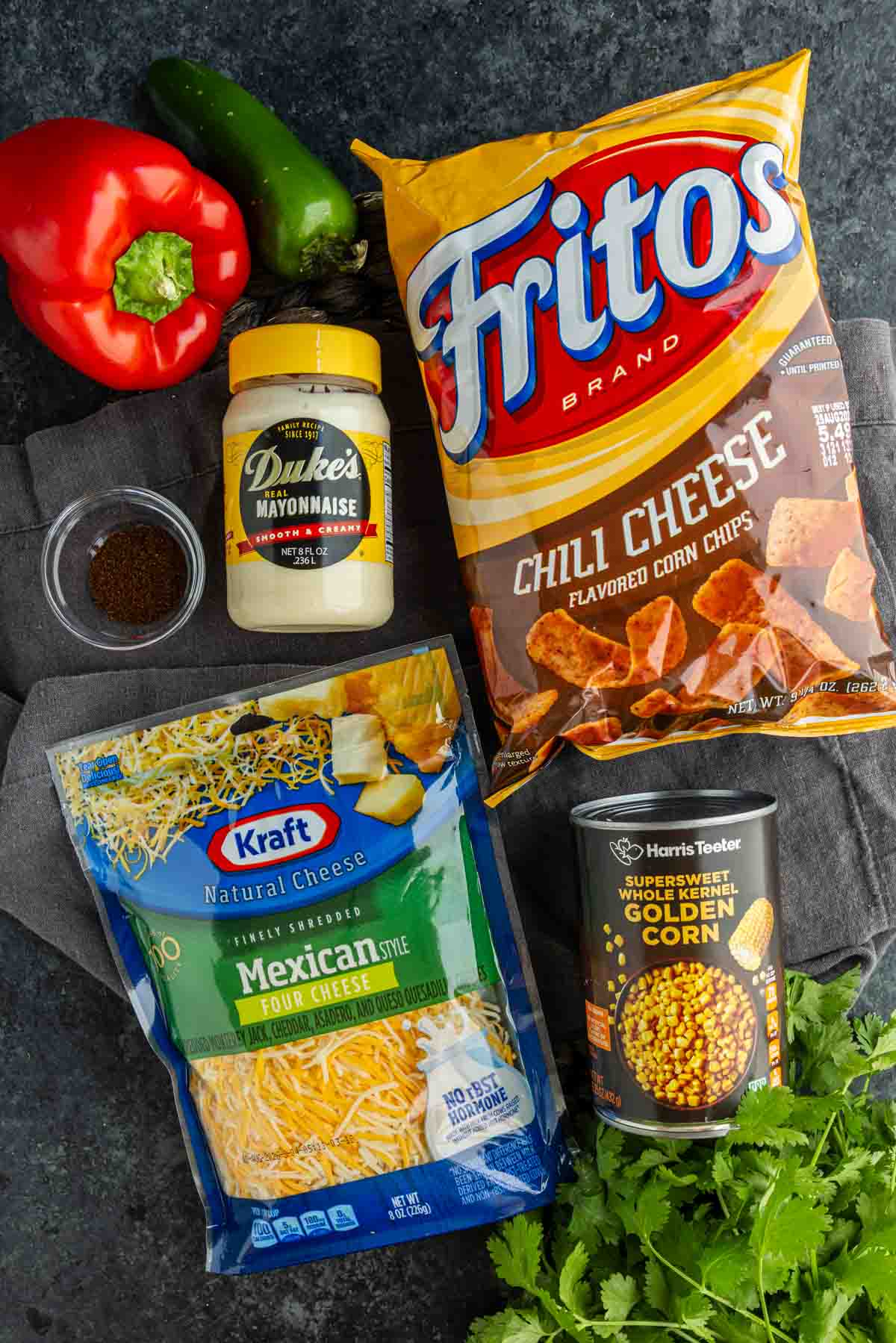 A bag of chili cheese Fritos, shredded Mexican cheese, canned corn, mayonnaise, chili powder, red bell pepper, jalapeño, and fresh cilantro displayed on a dark surface—everything you need for an easy Frito salad recipe.