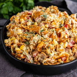 A black bowl filled with a creamy Frito Corn Salad, featuring corn, tomatoes, herbs, shredded cheese, and crunchy chips, placed on a dark cloth.