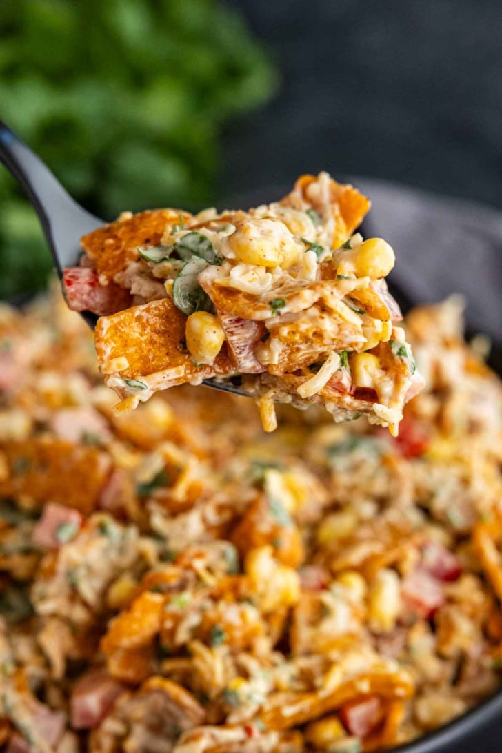 A close-up of a fork holding a portion of corn chip salad with cheese, corn, diced tomatoes, herbs, and tortilla chips above a bowl of the same easy Frito salad recipe.
