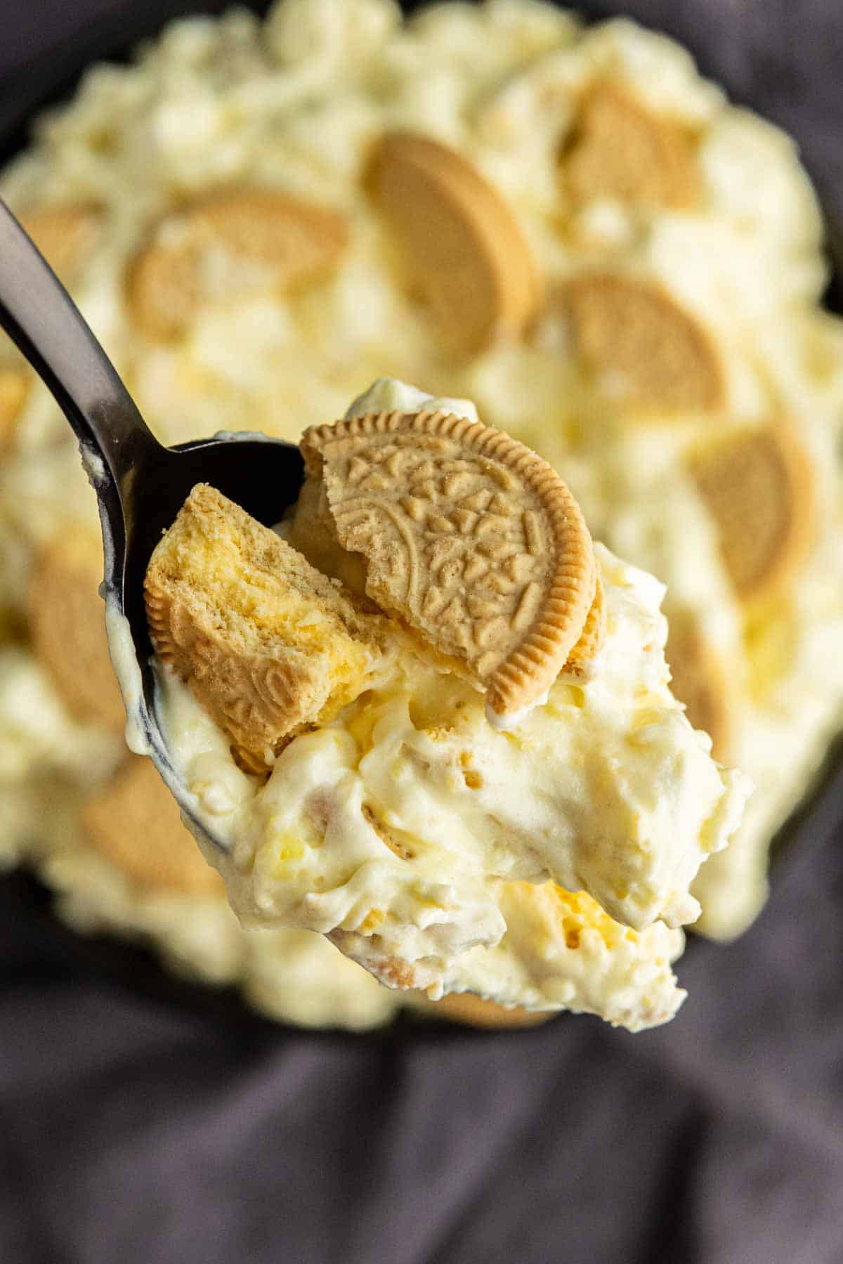 A close-up of a spoonful of creamy lemon fluff dessert with crushed golden sandwich cookies, held above a dish of the same lemon dessert.