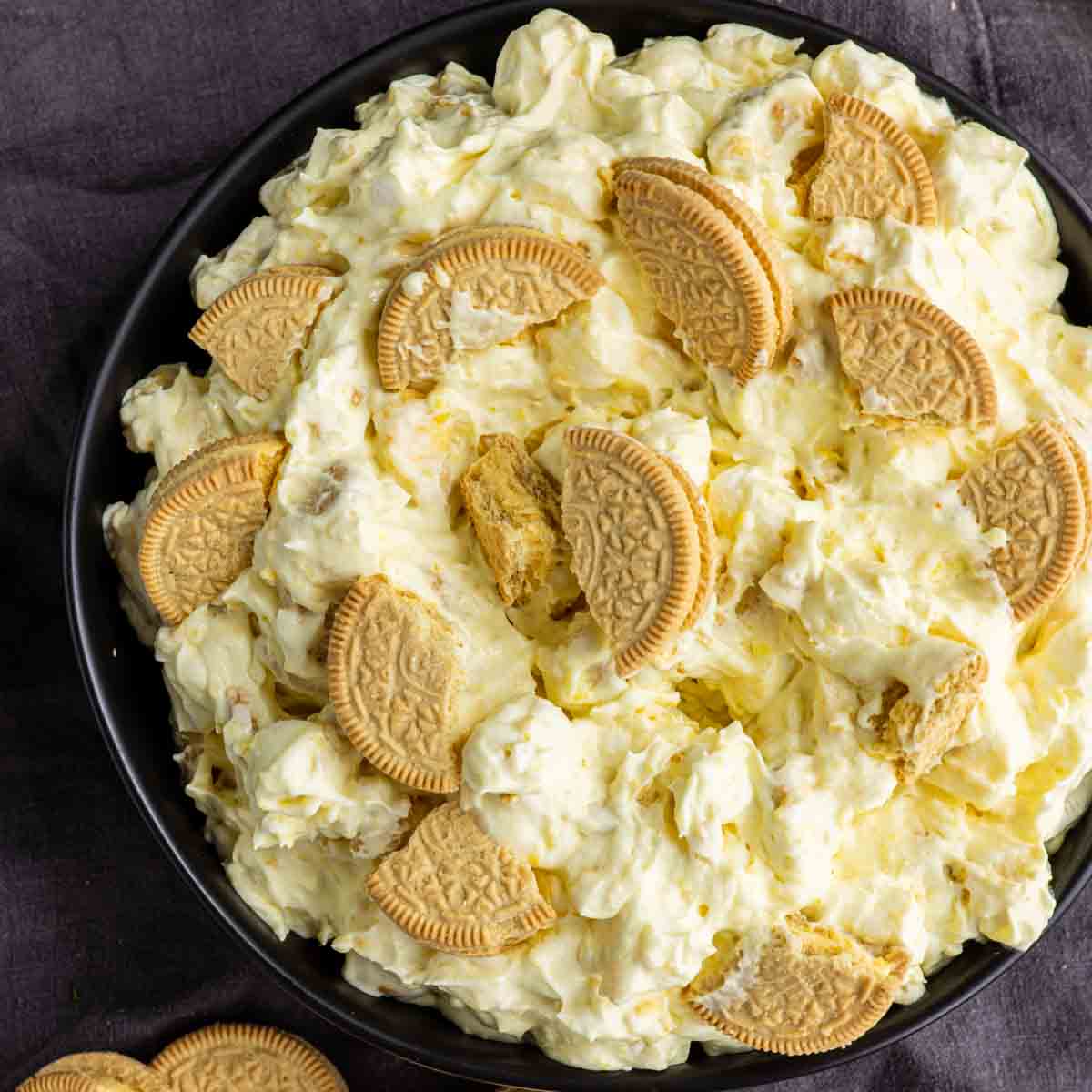 A bowl filled with a creamy lemon dessert salad topped with pieces of golden sandwich cookies, displayed on a dark surface.