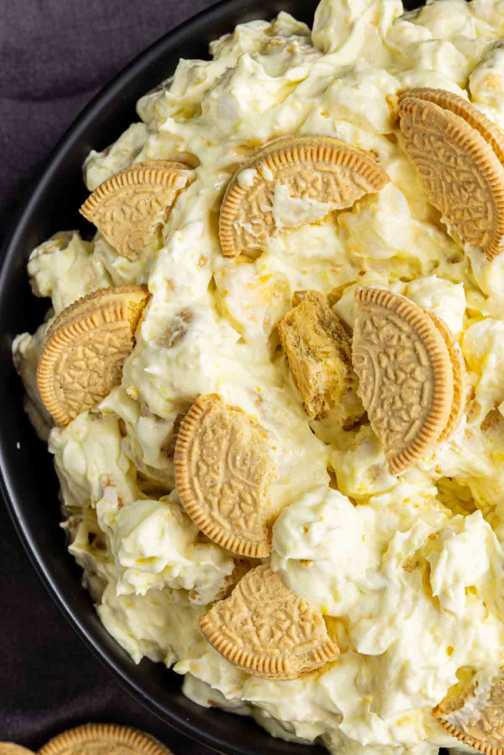 A close-up of a lemon dessert salad with a creamy, yellowish mixture and pieces of golden sandwich cookies on top, served in a black bowl—an irresistible take on the classic lemon fluff.