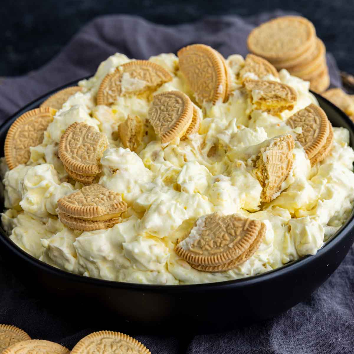 A black bowl filled with a creamy lemon fluff dessert mixture, topped with pieces of golden sandwich cookies, with more cookies scattered around the bowl.