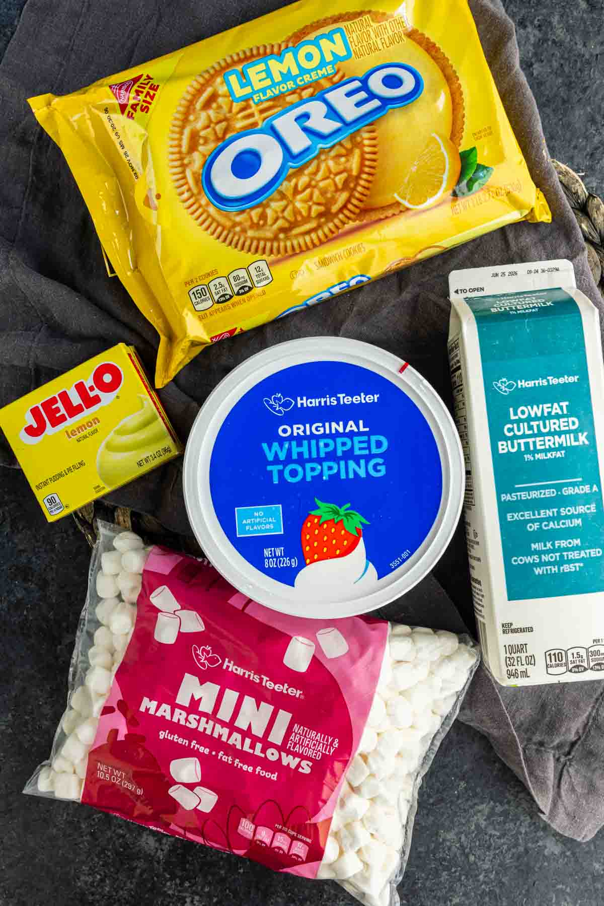 A carton of buttermilk, whipped topping, mini marshmallows, lemon Jello mix, and lemon Oreos are arranged on a dark surface—perfect for making a refreshing lemon fluff or your favorite lemon cookie salad recipe.