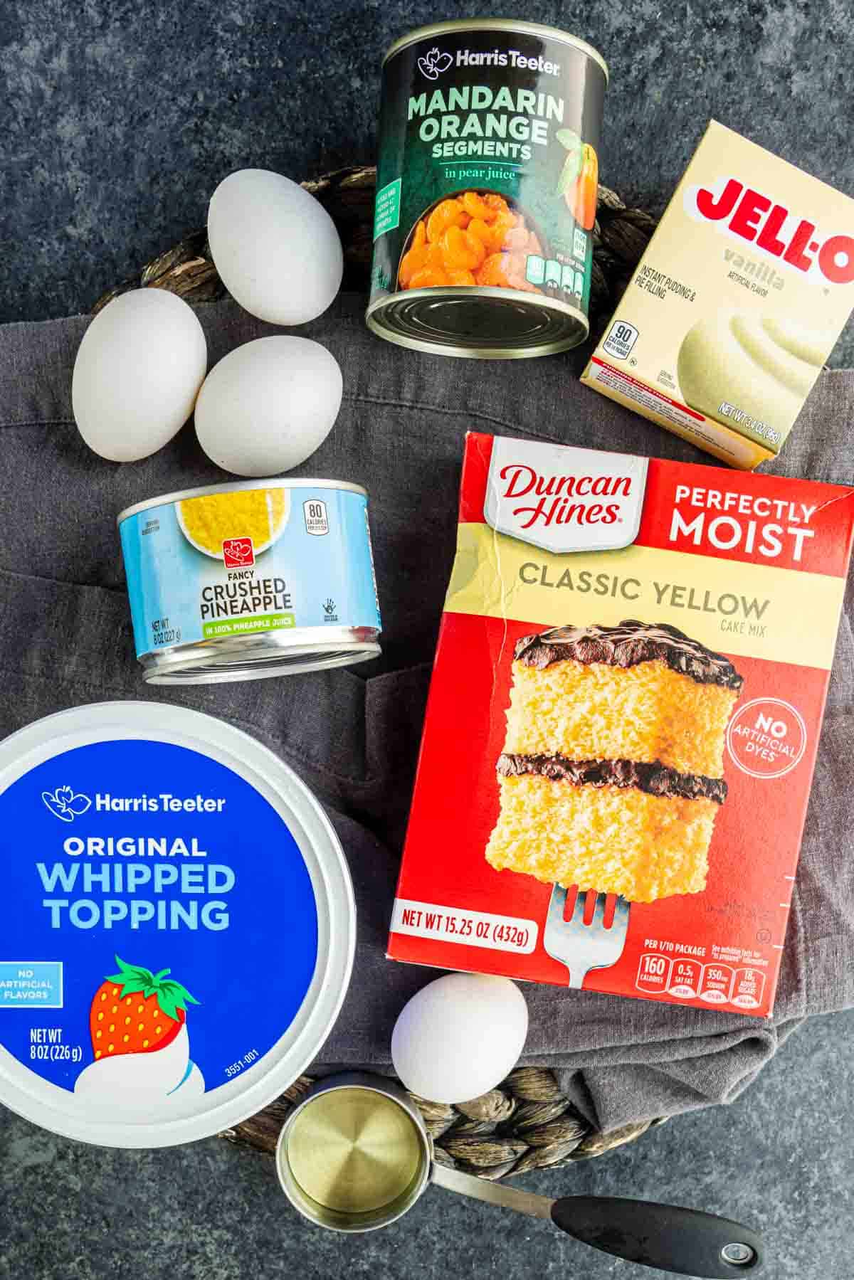 Ingredients for a Pig Pickin’ Cake, including eggs, whipped topping, crushed pineapple, mandarin oranges, vanilla pudding mix, and classic yellow cake mix, arranged on a dark surface.