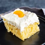 A square piece of mandarin orange cake with white whipped frosting, topped with a mandarin orange slice, on a black plate.