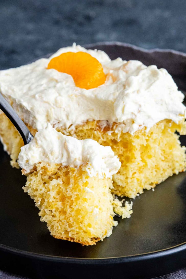 A slice of Pig Pickin’ Cake—a classic Southern dessert—topped with whipped cream and a mandarin orange segment, served on a black plate with a fork holding a bite.