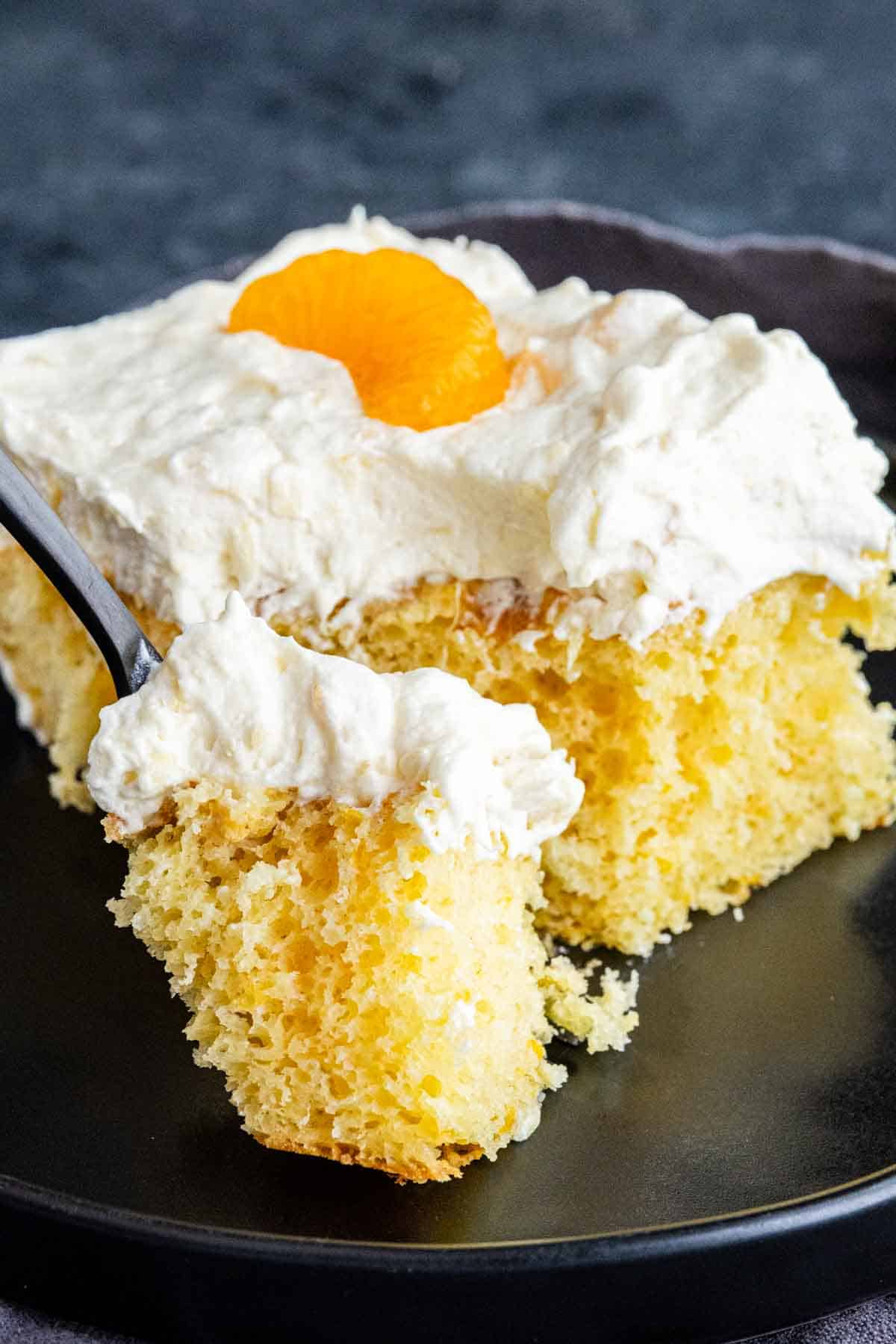 A slice of Pig Pickin’ Cake—a classic Southern dessert—topped with whipped cream and a mandarin orange segment, served on a black plate with a fork holding a bite.